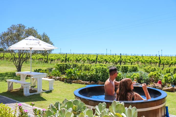Two guests relaxing in the wooden hot tub in the garden, with the vineyards and a picnic table behind