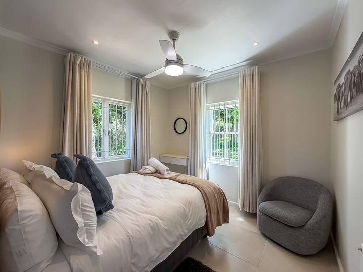A bedroom with a ceiling fan, a grey armchair and cream curtains over the garden windows