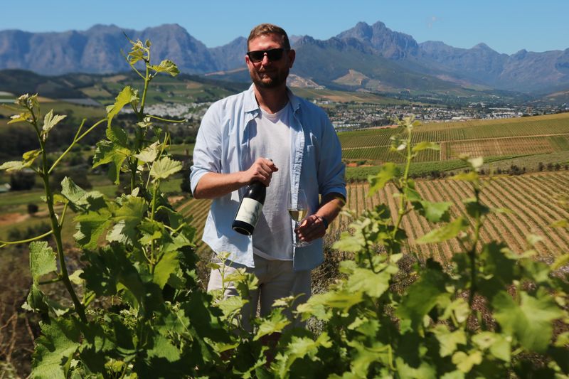A man holding a bottle of wine among the vines, with the mountains behind